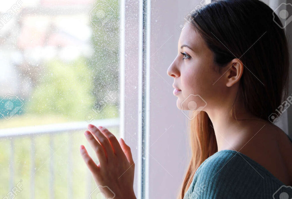 una fotografia de una persona mirando por una ventana con expresion melancolica