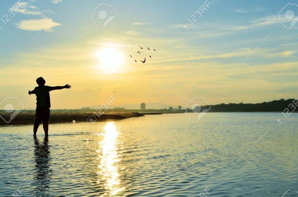 una imagen con una puesta de sol sobre un paisaje sereno y una persona con las manos levantadas en adoracion