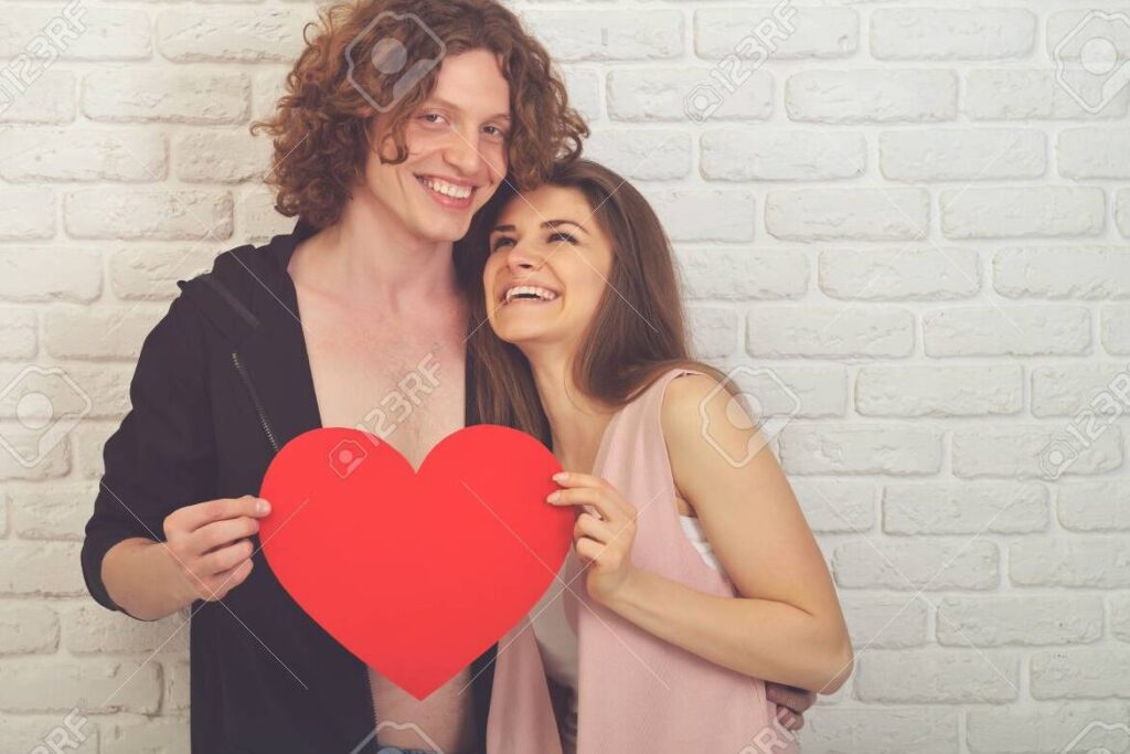 una pareja joven se abraza y sonrie frente a un fondo de corazones rojos