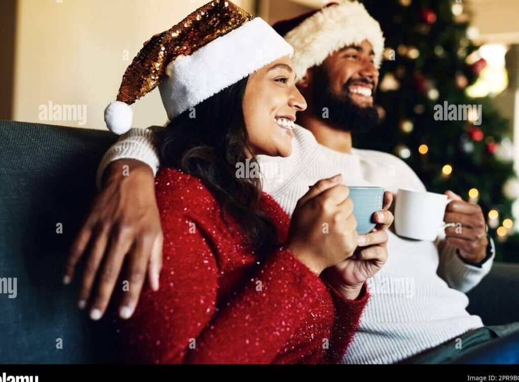 una pareja sonriente sosteniendo una taza de chocolate caliente con malvaviscos frente a un arbol de navidad decorado