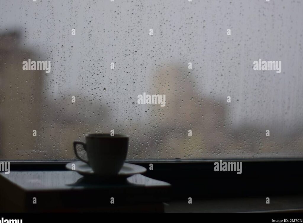 una taza de cafe humeante sobre una mesa de madera con una ventana de fondo mostrando la lluvia cayendo suavemente afuera